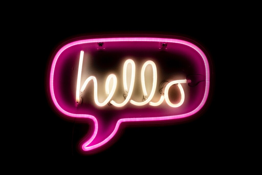Leuchtender &bdquo;hello&ldquo;-Schriftzug in einer pinken Neon-Sprechblase vor schwarzem Hintergrund.